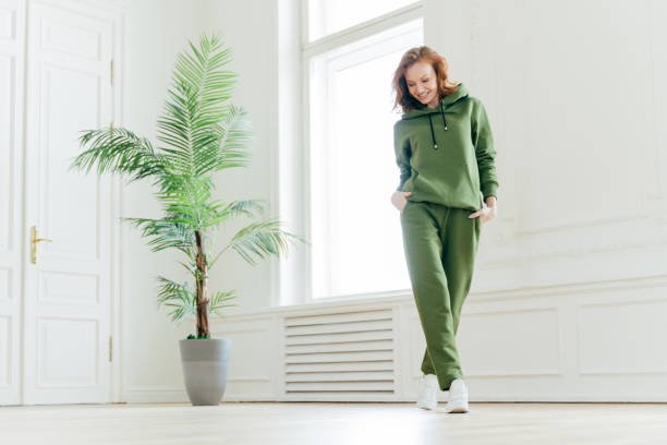Full length shot of active pleased young woman with slim body, keeps hands in pockets of tracksuit, focused down, ready for doing cardio exercises, has determined look, pilates exercising indoor