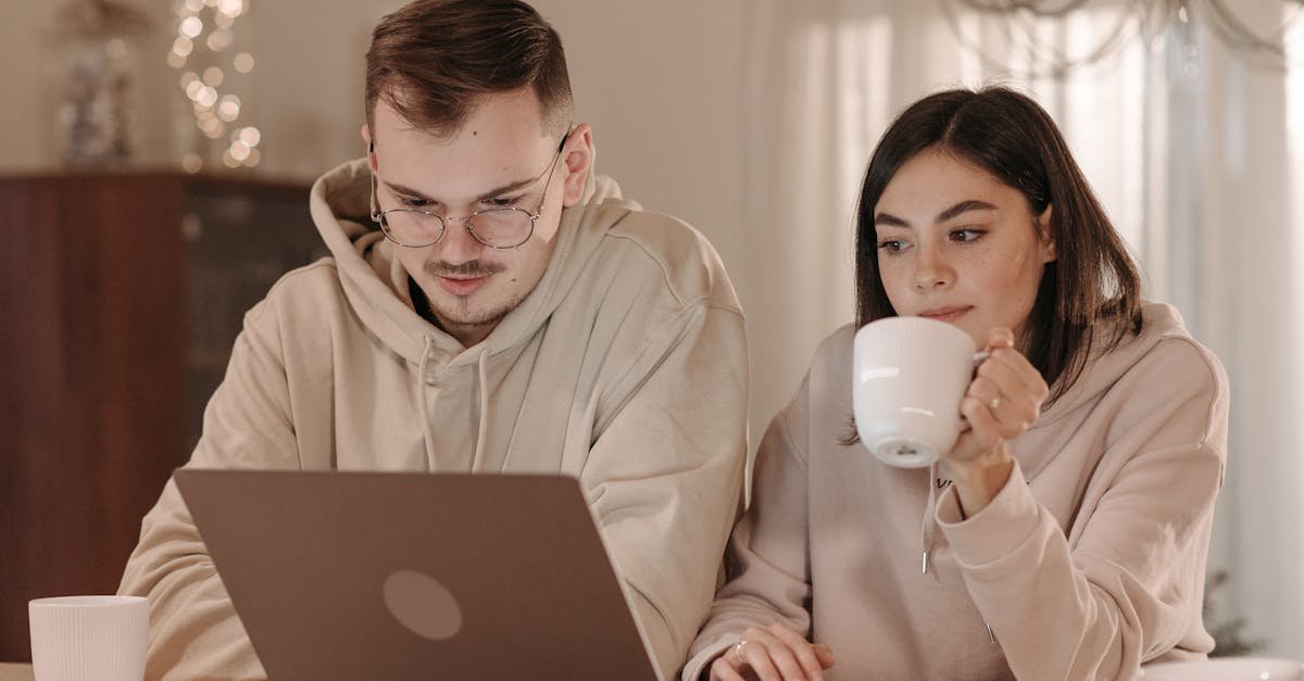A couple in casual hoodies working from home with coffee and a laptop.